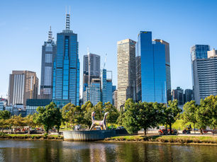 City skyline with tall blue and beige skyscrapers under a clear sky, trees in the foreground, and a reflective river below. Vibrant and calm.