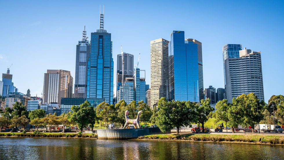 City skyline with tall blue and beige skyscrapers under a clear sky, trees in the foreground, and a reflective river below. Vibrant and calm.