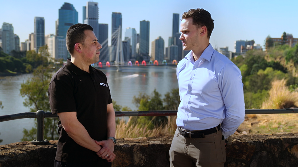 Two real estate professionals in conversation with the Brisbane city skyline in the background.
