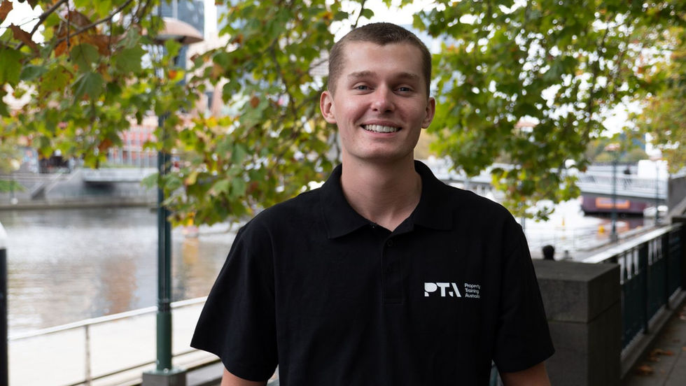 Smiling man in black PTA polo stands by a riverside walkway under green trees.